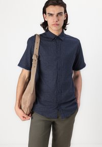 Kurzärmeliges marineblaues Hemd mit subtilem Muster, Knopfleiste, kombiniert mit olivgrünen Hosen und einer beigen Umhängetasche.