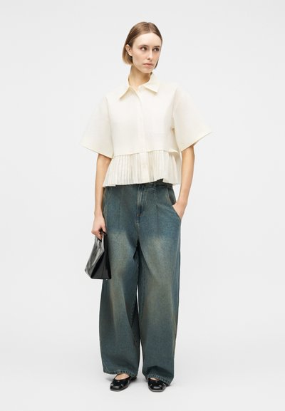 Γυναίκα που φοράει λευκό μπλουζάκι με κοντά μανίκια και πιέτες στο τελείωμα, φαρδιά ξεβαμμένα τζιν, μαύρα φλατ παπούτσια και κρατάει μια μικρή μαύρη τσάντα.