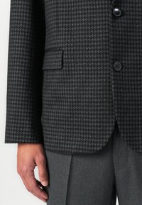 Männliche Hand neben einem dunkelgrauen Hahnentrittmuster-Blazer mit schwarzen Knöpfen und passenden dunkelgrauen gefaltenen Hosen.