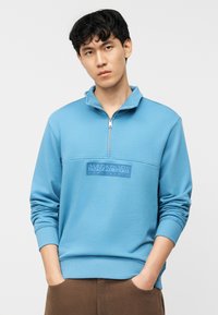 Νέος άνδρας που φοράει μπλε φούτερ με φερμουάρ στο λαιμό και το λογότυπο "Napapijri geographic", με τα χέρια στις τσέπες καφέ παντελονιού, μπροστά σε απλό λευκό φόντο.