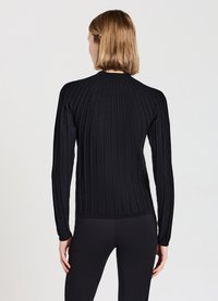 Frau mit schulterlangen blonden Haaren, die einen schwarzen gerippten Langarmpullover und schwarze eng anliegende Hosen trägt, mit dem Rücken zu einer schlichten Hintergrund.