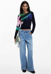 Námornícky modrý top s dlhými rukávmi a farebnou kvetinovou potlačou, kombinovaný s svetlomodrými širokými džínsami. Doplnok tvorí malá biela kabelka.