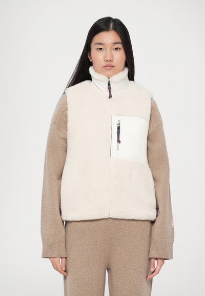 Kvinde med langt mørkt hår iført en beige striktrøje og bukser samt en ærmeløs hvid fleecevest med høj krave og lynlås-lomme.