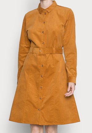Femme portant une robe camel à manches longues, boutonnée, avec col et ceinture assortie à la taille.