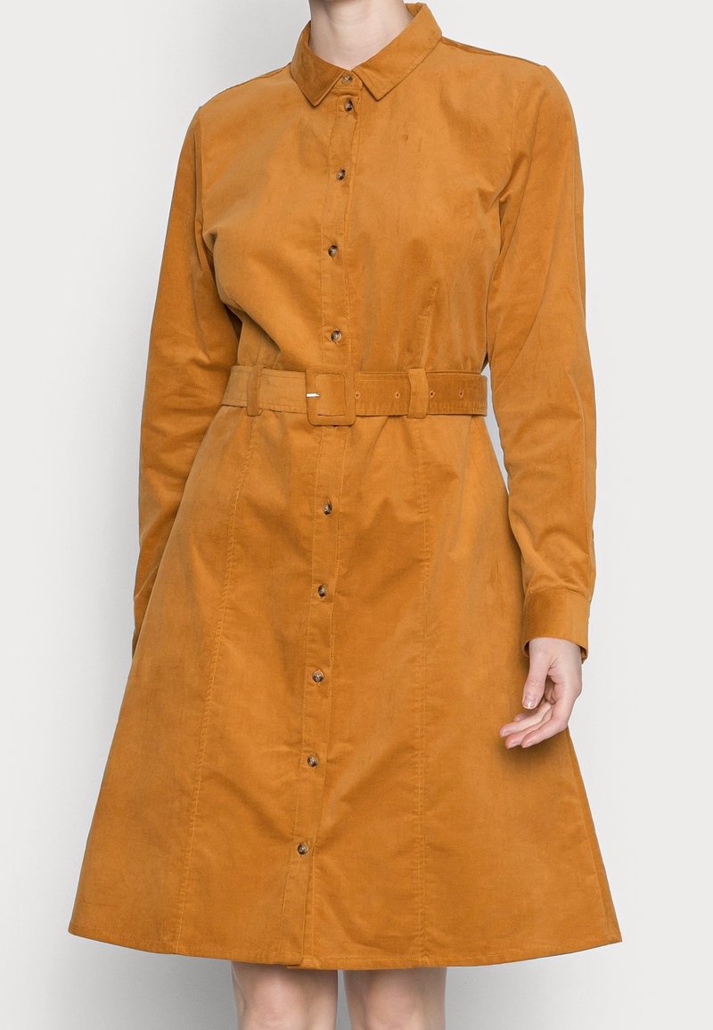 Femme portant une robe camel à manches longues, boutonnée, avec col et ceinture assortie à la taille.
