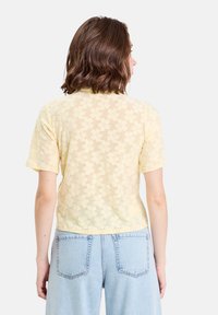 Haut à manches courtes en dentelle florale jaune avec un design transparent. Présente un col et une coupe ajustée, rehaussée d'un ourlet arrondi.