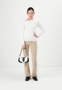Young woman in white long-sleeve top, beige corduroy pants, black shoes, holding a small black-and-white handbag, standing against a white background.