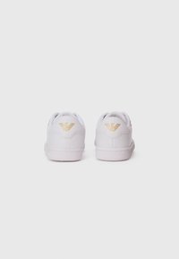 Et par hvite sneakers sett bakfra, med gull-logo detaljer på hælen mot en enkel hvit bakgrunn.