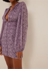 Robe mini en dentelle florale lavande avec de longues manches bouffantes, décolleté en V profond, boutons à l'avant, et détails froncés aux poignets et à la taille.