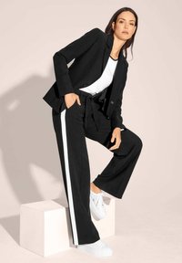 Schwarze Blazer und weite Hosen mit weißen Seitenstreifen, kombiniert mit einem weißen T-Shirt und weißen Sneakers, vor einem neutralen Hintergrund.