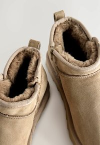 Botines de ante beige con forro de piel suave, dos lengüetas y suela texturizada. Detalle de cremallera visible en el costado.