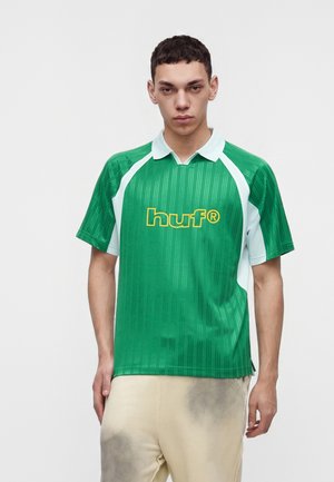 Ung mand iført en grøn og hvid sportsbluse med krave og gul "huf"-logo, kombineret med lyse beige bukser.
