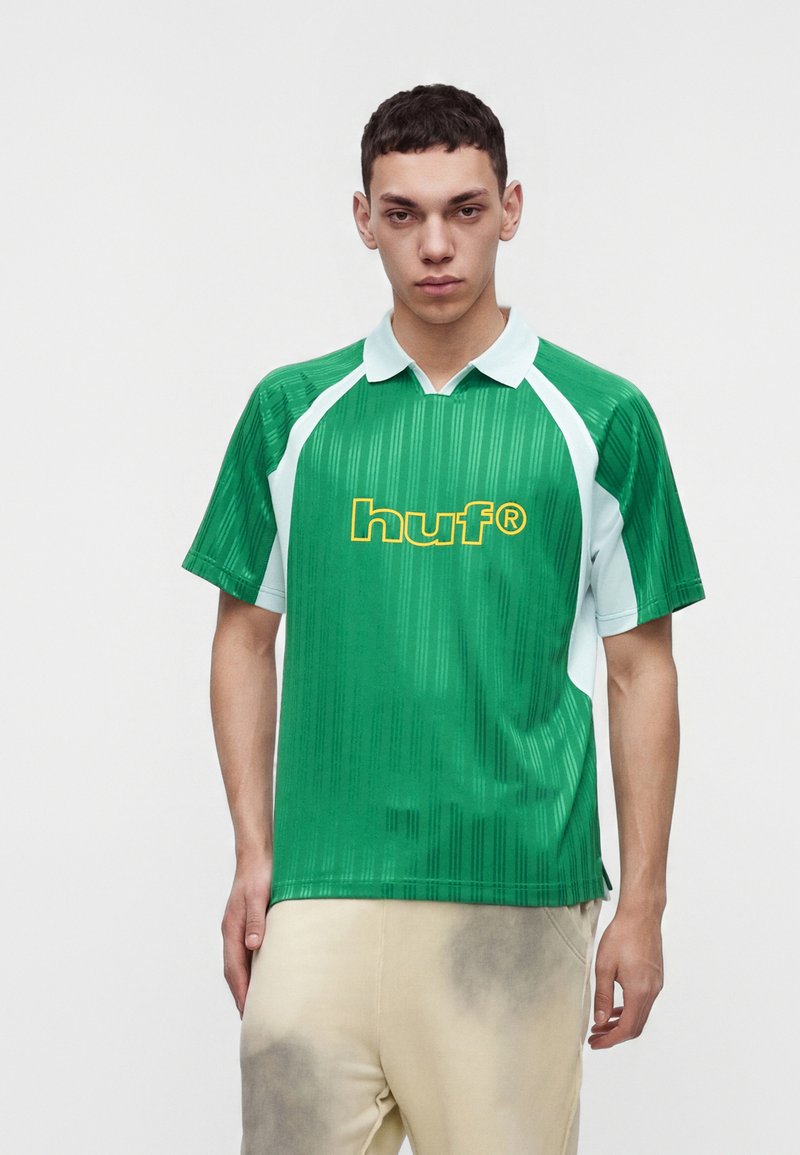 Jeune homme portant un polo de sport vert et blanc avec le logo « huf » en jaune, assorti à un pantalon beige clair.
