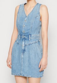 Denimklänning med v-ringning, ärmlös design, silverfärgad knäppning, definierad midja och två framfickor. Ljusblå färg och blekt utseende.