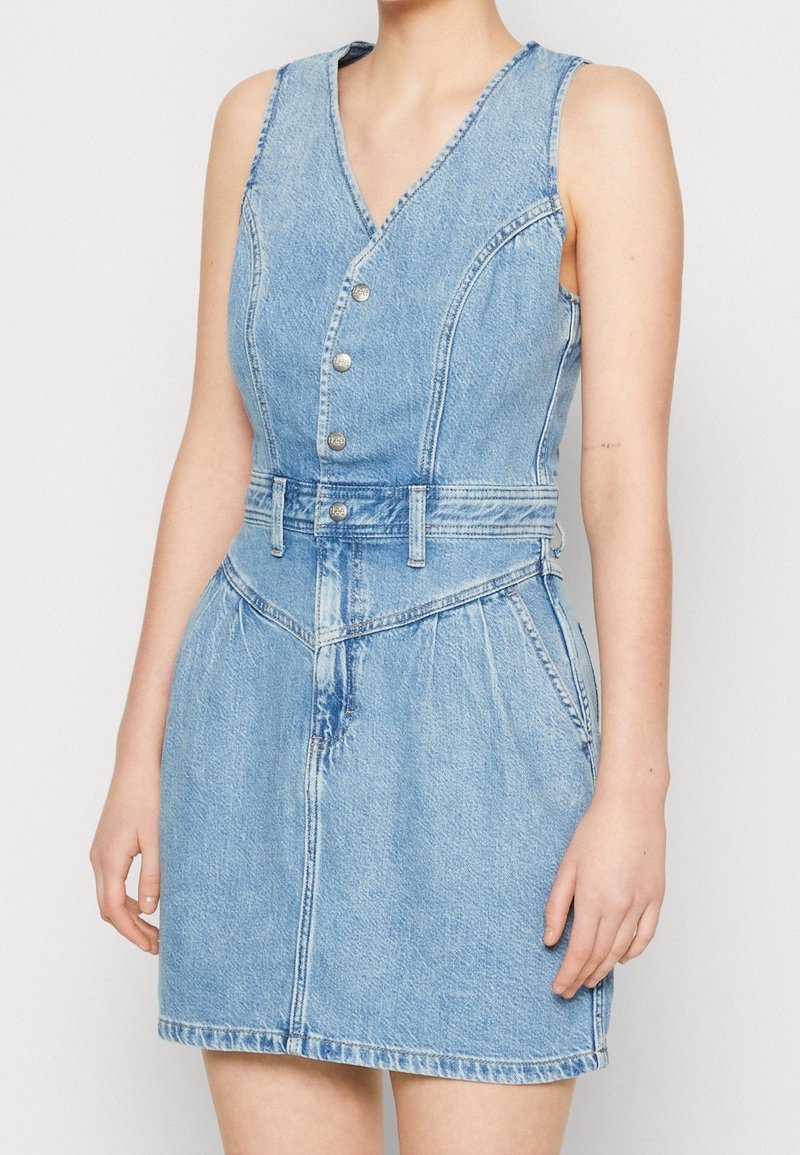 Denimklänning med v-ringning, ärmlös design, silverfärgad knäppning, definierad midja och två framfickor. Ljusblå färg och blekt utseende.