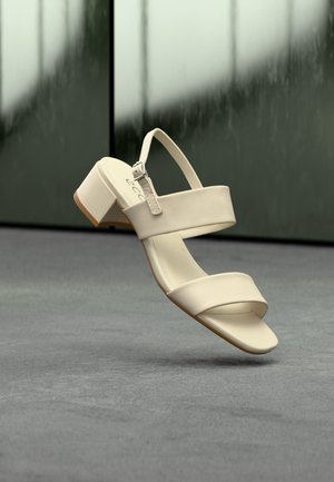 Smėlio spalvos atviros kojų pirštų basutės su dviem dirželiais ir žemu kubo formos kulnu, pavaizduotos kabant virš pilko paviršiaus prie neaiškios tamsios fono.