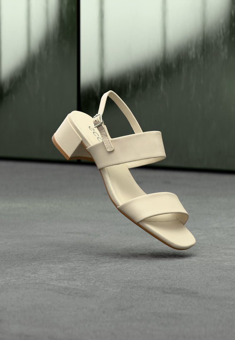 Sandale beige à bout ouvert avec deux brides et un petit talon bloc, suspendue au-dessus d'une surface grise sur un fond sombre flou.