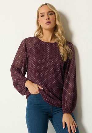 Kvinde med langt blond hår har en gennemsigtig vinrød bluse med hvide prikker og blå jeans på, stående mod en ensfarvet lys baggrund.