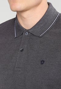 Grå pikétröja med en strukturerad tyg, med en treknapps stängning, en krage med vit kant och ett litet emblem på bröstet.