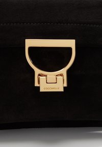 Gros plan d’un sac à main en daim noir avec un fermoir en métal doré gravé du nom de la marque "Coccinelle".