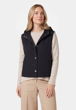 Frau trägt eine schwarze ärmellose Kapuzenweste über einem beigen Langarmpullover und beigen Hosen, steht mit entspannt an den Seiten hängenden Händen.