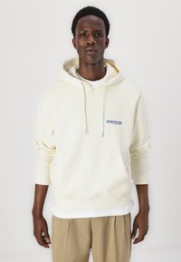 Un hoodie couleur crème en tissu doux, avec une poche kangourou à l'avant et des cordons de serrage, portant un logo bleu sur la poitrine gauche.