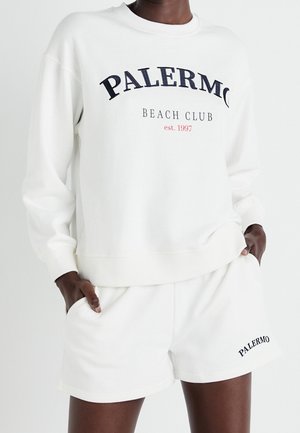 Personne portant un sweat-shirt blanc et un short avec un texte noir "PALERMO BEACH CLUB est. 1997" sur le sweat-shirt et "PALERMO" sur le short.