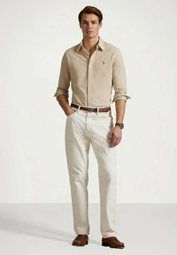 Beige langermet skjorte med knapper, lysfargede bukser, brun lærbelte og sko, med en liten logo på skjorten.