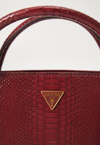 Sac à main en cuir rouge texturé avec double poignée et logo triangulaire doré Guess USA au centre devant.