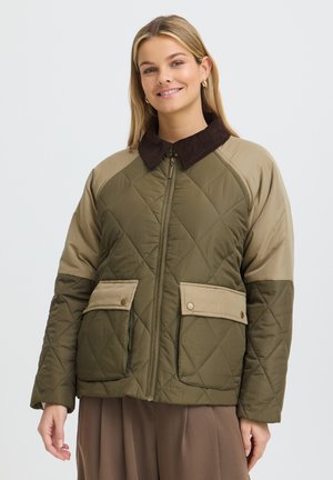 Femme souriante, portant une veste matelassée vert olive avec des manches et poches beige, et un col foncé, debout devant un fond uni.