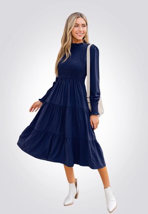 Vestido azul marino de capas con corsé fruncido y mangas largas, hecho de una tela de textura suave. Llevado con botas blancas hasta el tobillo.