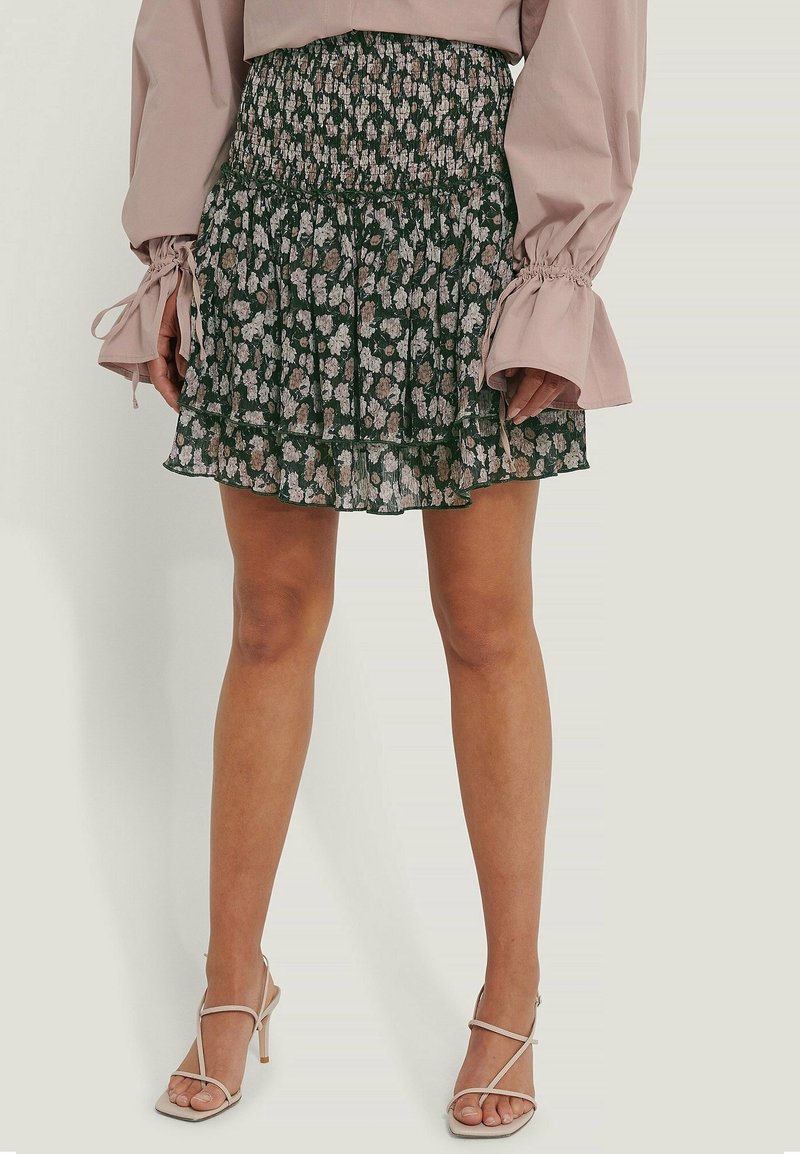 Falda floral verde con cintura fruncida y volantes en capas; presenta pequeñas flores blancas y beige. Combinada con tacones beiges con tiras.