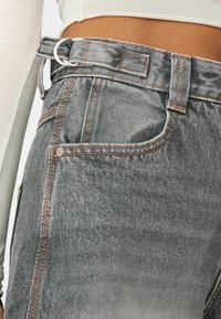 Zone de la taille et des hanches d'une personne portant un jean gris taille haute avec des coutures apparentes et une ceinture en tissu avec une boucle double anneau argentée.