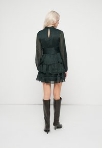 Σκούρο πράσινο φόρεμα με στ�ρώσεις, ψηλό λαιμό, διάφανες μακριές μανίκες και λεπτομέρεια με άνοιγμα στην πλάτη, συνδυασμένο με καφέ ψηλές γόβες.