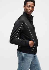 Μαύρο μπουφάν με φερμουάρ και λευκές λωρίδες κατά μήκος των ραφών, κατασκευασμένο από λείο ύφασμα. Το μοντέλο το φορά πάνω από ανοιχτόχρωμα μπλε τζιν, με τα χέρια στις τσέπες.