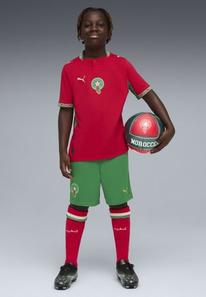 Jongere staat met een voetbal met Marokkaans thema, draagt het rood-groene Marokkaanse voetbaluniform en zwarte voetbalschoenen tegen een grijze achtergrond.