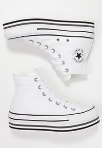 Baskets montantes blanches en toile, avec des œillets argentés, des semelles en caoutchouc noires ornées de détails rayés blancs et noirs, et un logo étoile sur le côté.