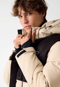 Beige en zwarte gewatteerde jas met een hoge kraag, voorzien van een rits en verstelbare capuchon, een gladde textuur en contrasterende details op de mouwen.