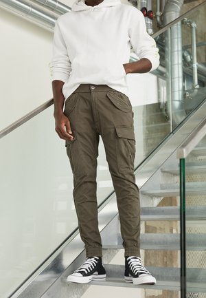 Persoon met een witte hoodie, olijfkleurige cargobroek en zwarte hoge sneakers, staand op metalen trappen binnen met glazen leuning en zichtbare leidingen.