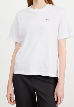 Witte T-shirt met korte mouwen en ronde hals, gedragen door een persoon, met een klein groen krokodillenlogo op de linkerborst, gecombineerd met een zwarte broek.