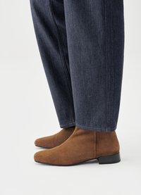 Bottines en daim marron avec un bout pointu et un talon noir bas, portées avec un pantalon en denim foncé à coupe droite et ourlet légèrement effiloché.