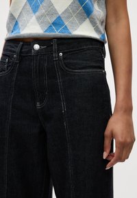 Črne denim hlače z kontrastnim šivanjem, prilegajočim pasom in ravnim krojem. Roka je postavljena ob stranski šiv.