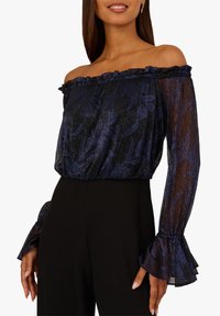 Top bez ramion z przezroczystej czarnej tkaniny z niebieskim wzorem kwiatowym. Zawiera falbaniasty dekolt oraz szerokie, warstwowe rękawy.