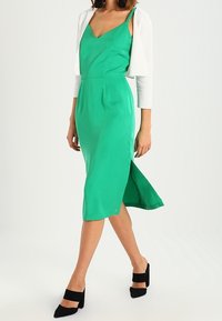 Zelené midi šaty bez rukávů s výstřihem do V, bočním rozparkem a hladkou texturou, doplněné bílým kardiganem a černými botami s masivním podpatkem.
