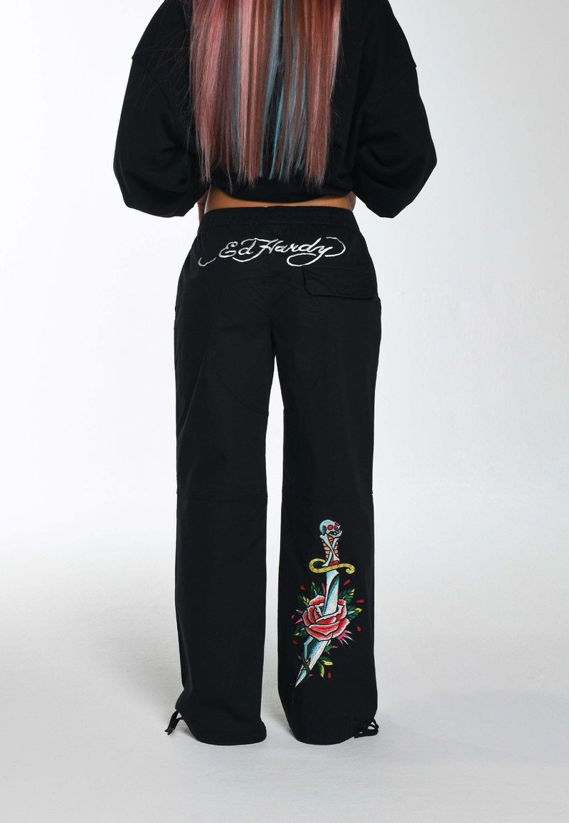 Black pants made from durable fabric feature vibrant embroidered designs of a dagger and rose, with a logo on the back waistband.