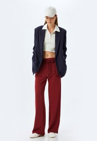Marineblazer med to knapper, hvid cropped polo, bordeaux brede sweatpants med snor, og hvide sneakers, toppet med en lys cap.