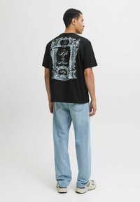 Czarna bawełniana koszulka z graficznym wzorem na plecach, z ozdobnymi ramkami i napisem "Życie jest teraz" na białym tle. Połączona z jasnoniebieskimi dżinsami.