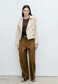 Beige kort jacka med dubbelknäppt design över en brun topp, som matchas med hög midja brun byxor och svarta sandaler.