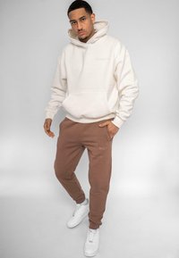 Hvid hættetrøje med frontlomme, parret med brune sweatpants med sidelommer. Afsluttet med hvide sneakers.