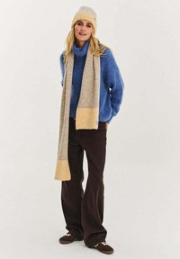 Modrý měkký svetr, béžovožlutý šátek, tmavě hnědé kalhoty s širokými nohavicemi, šedý klobouk a hnědé boty se světlými detaily. Neformální outfit.